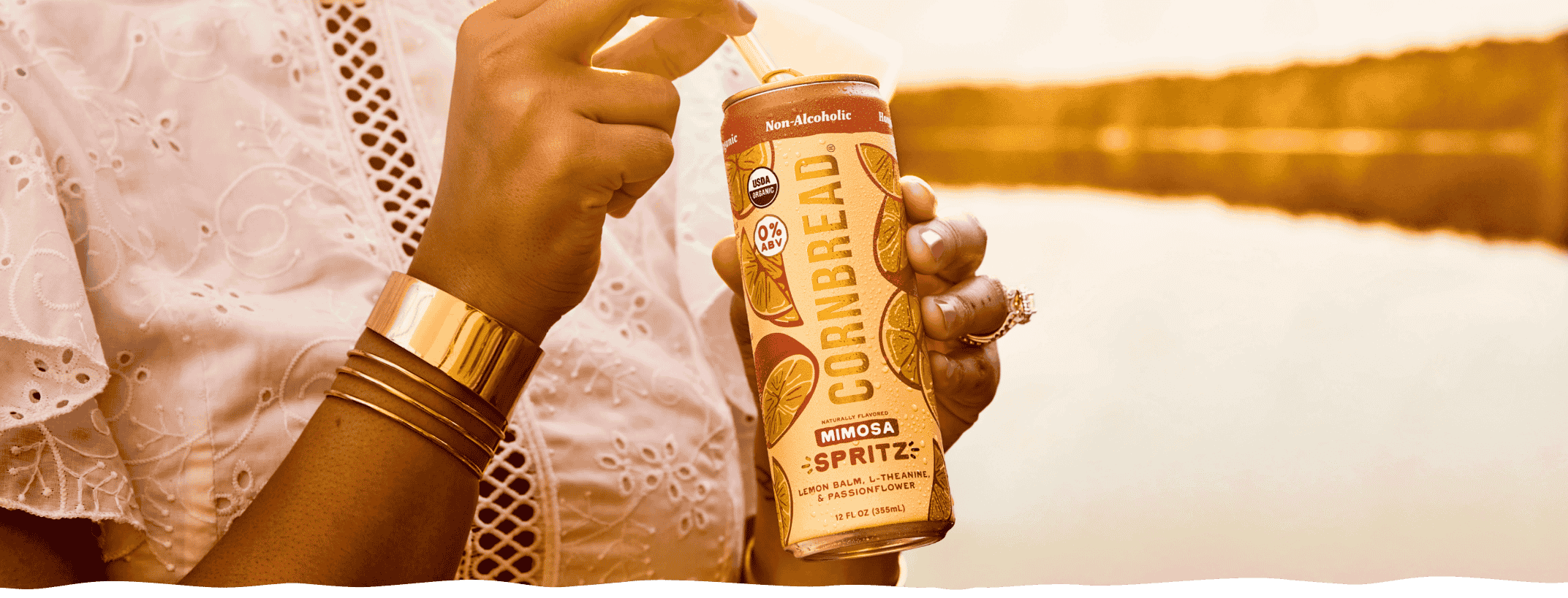 A woman holding a can of Mimosa Cornbread Social Spritz at golden hour by the lake